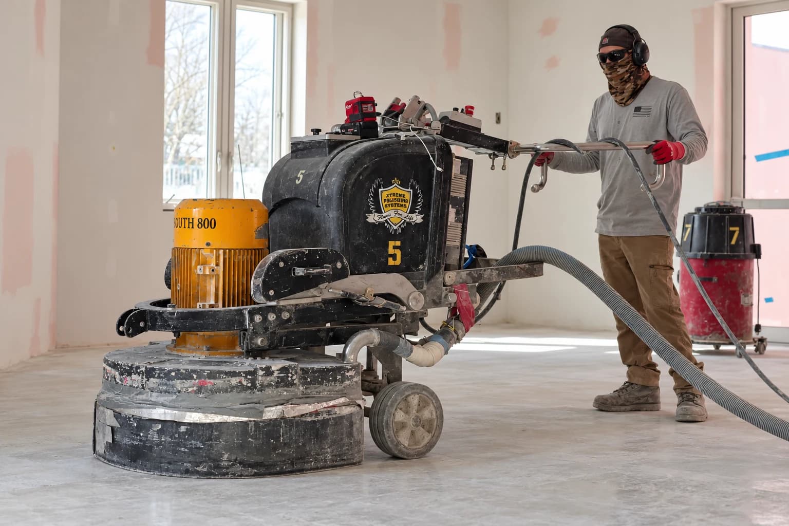An NCP crew member guiding a large planetary floor grinder across a slab mid-refine, daylight through tall windows