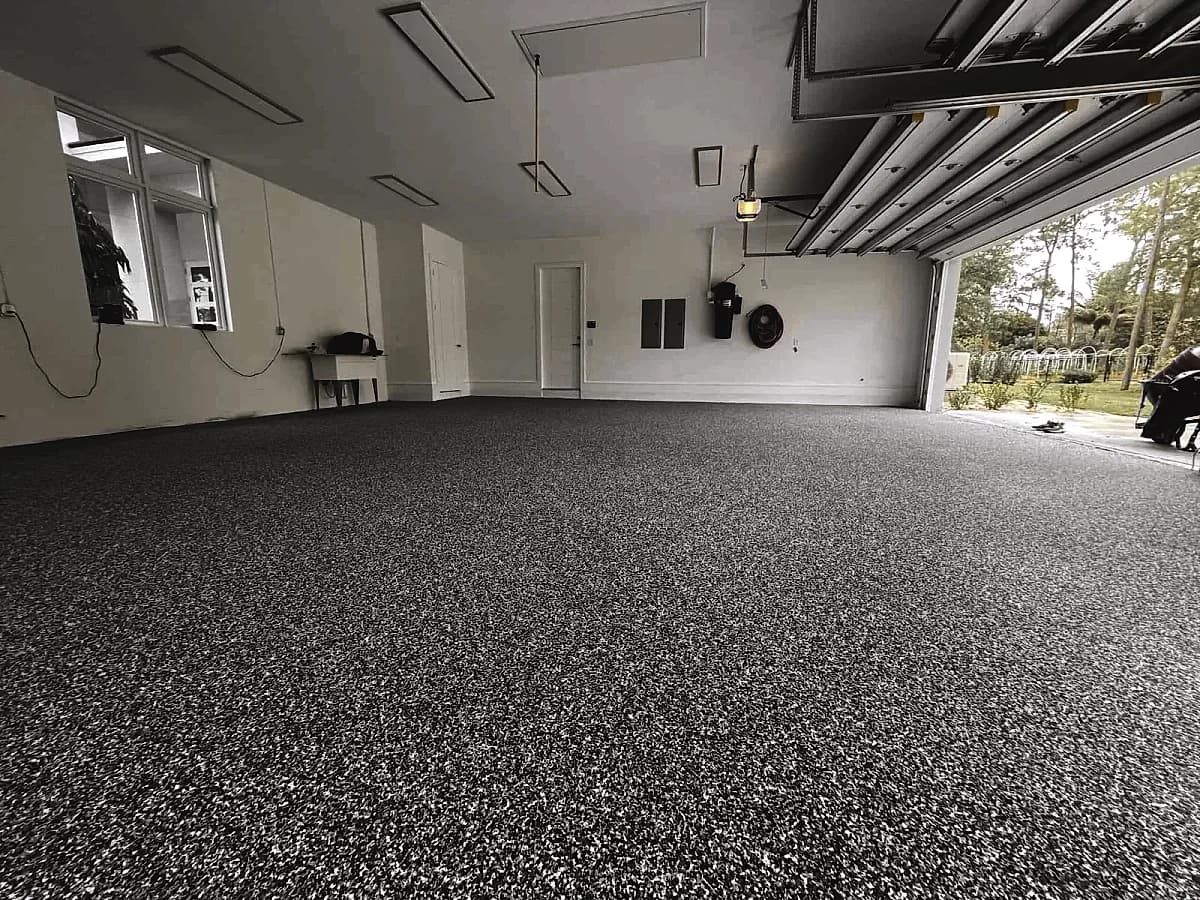 A residential garage with a gray flake floor, the overhead door open to the garden outside