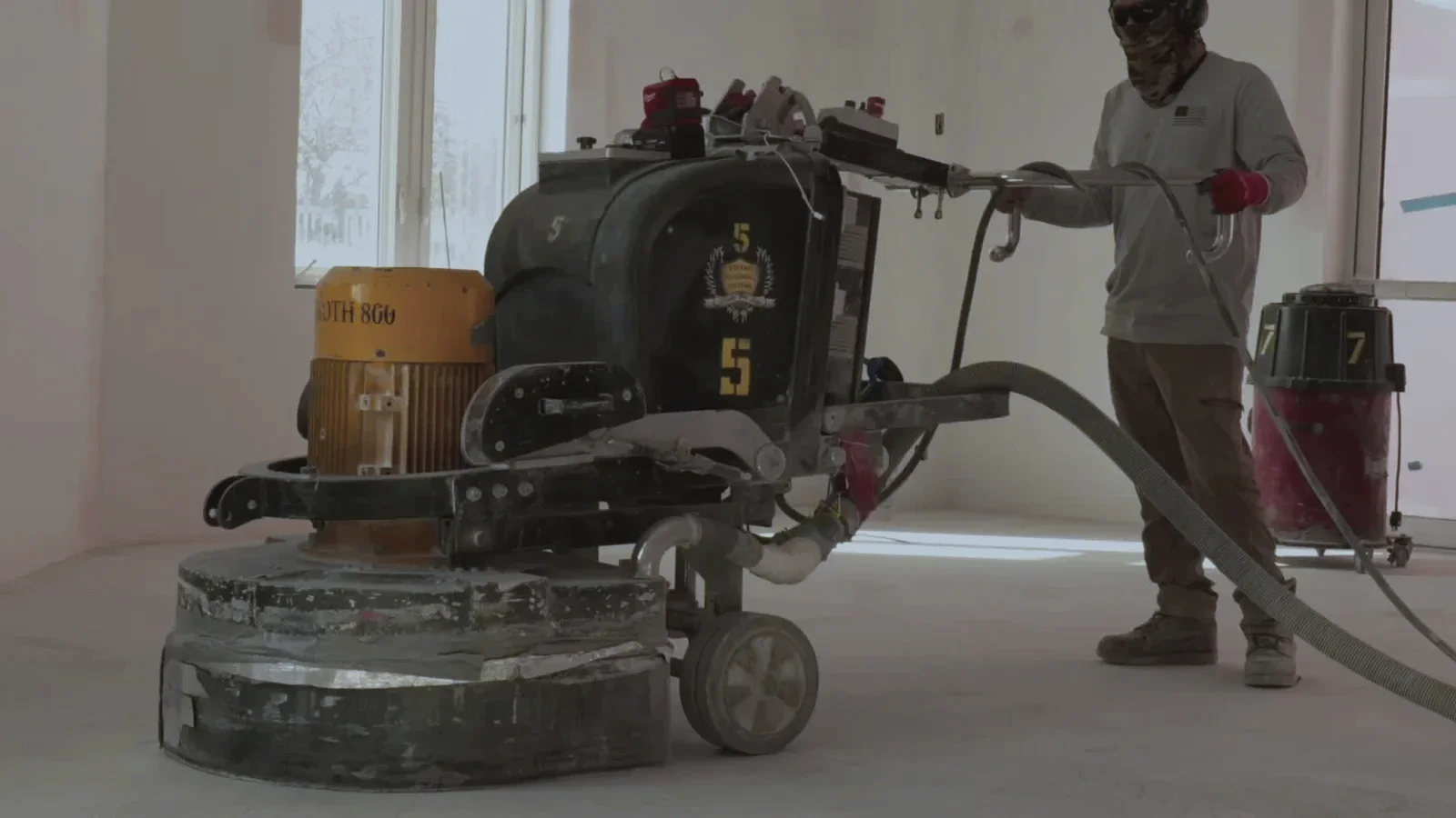 An NCP crew member guiding a large planetary floor grinder across a slab mid-refine, daylight through tall windows