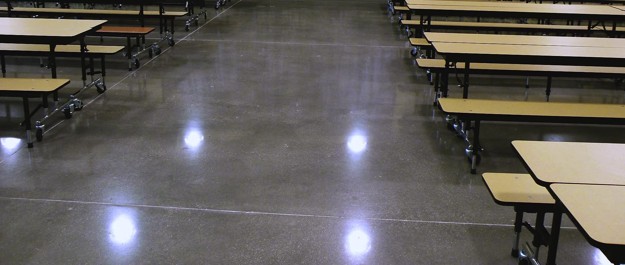 A school cafeteria with a ground and polished concrete floor that mirrors the ceiling lights in bright pools, rows of folding tables standing on it to either side