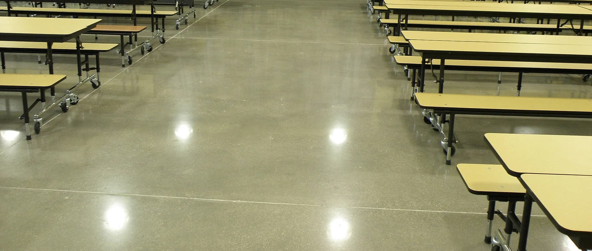 A school cafeteria with a ground and polished concrete floor that mirrors the ceiling lights in bright pools, rows of folding tables standing on it to either side