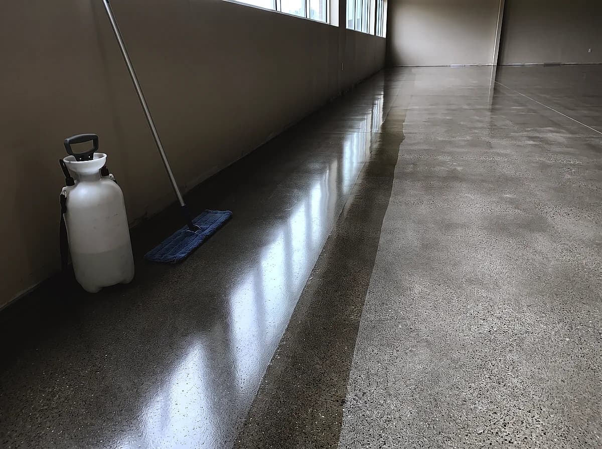 A polished concrete floor with daylight running down it as a long soft reflection, a pump sprayer and a pad on a handle at the wall