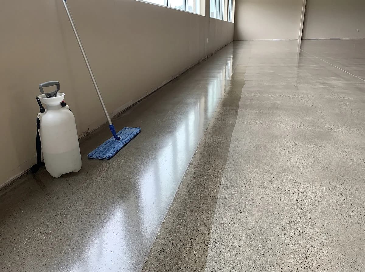A polished concrete floor with daylight running down it as a long soft reflection, a pump sprayer and a pad on a handle at the wall