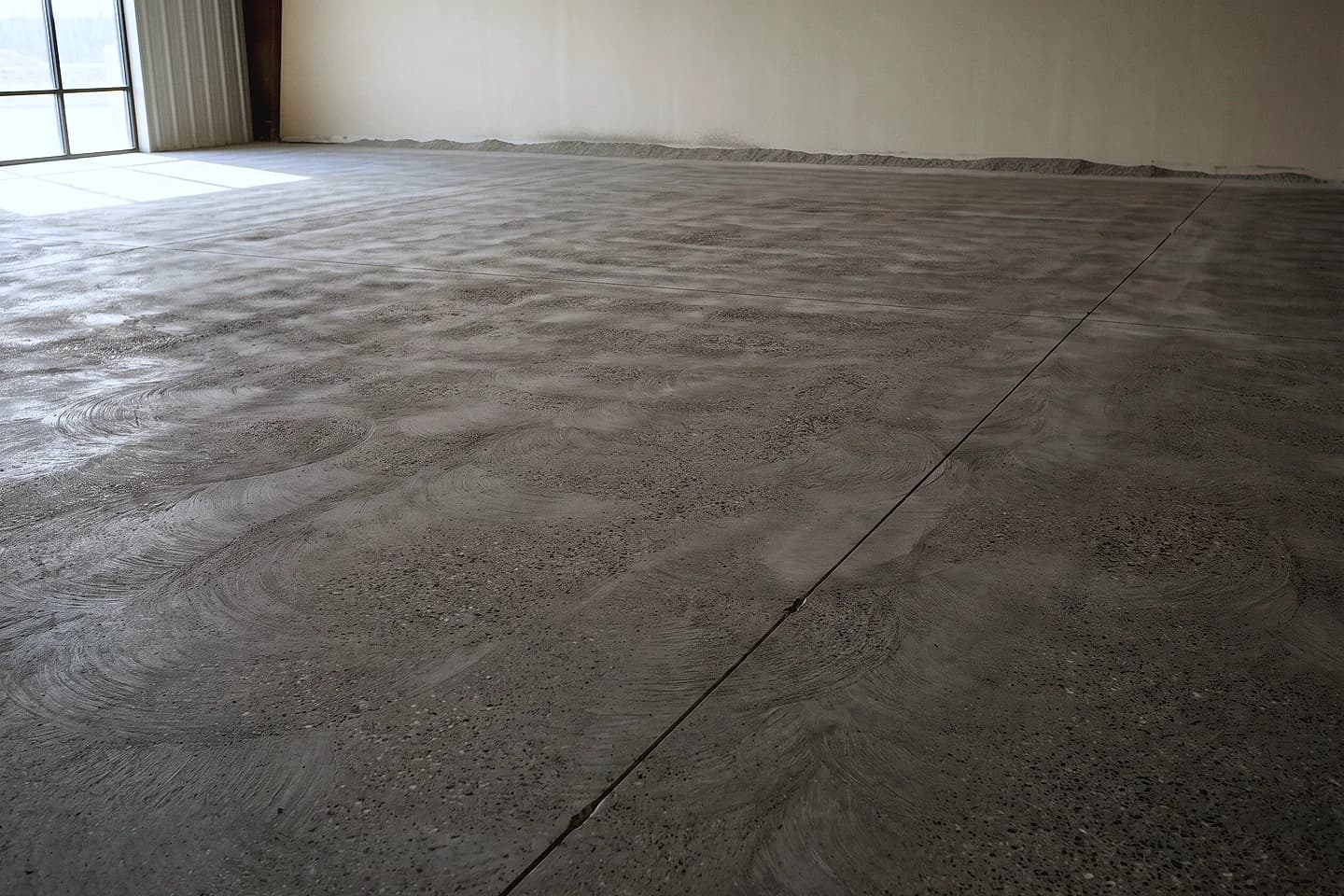 A bare concrete slab just after grinding, the grinder's overlapping arcs still visible across the open matte surface and a saw-cut joint running away to one side