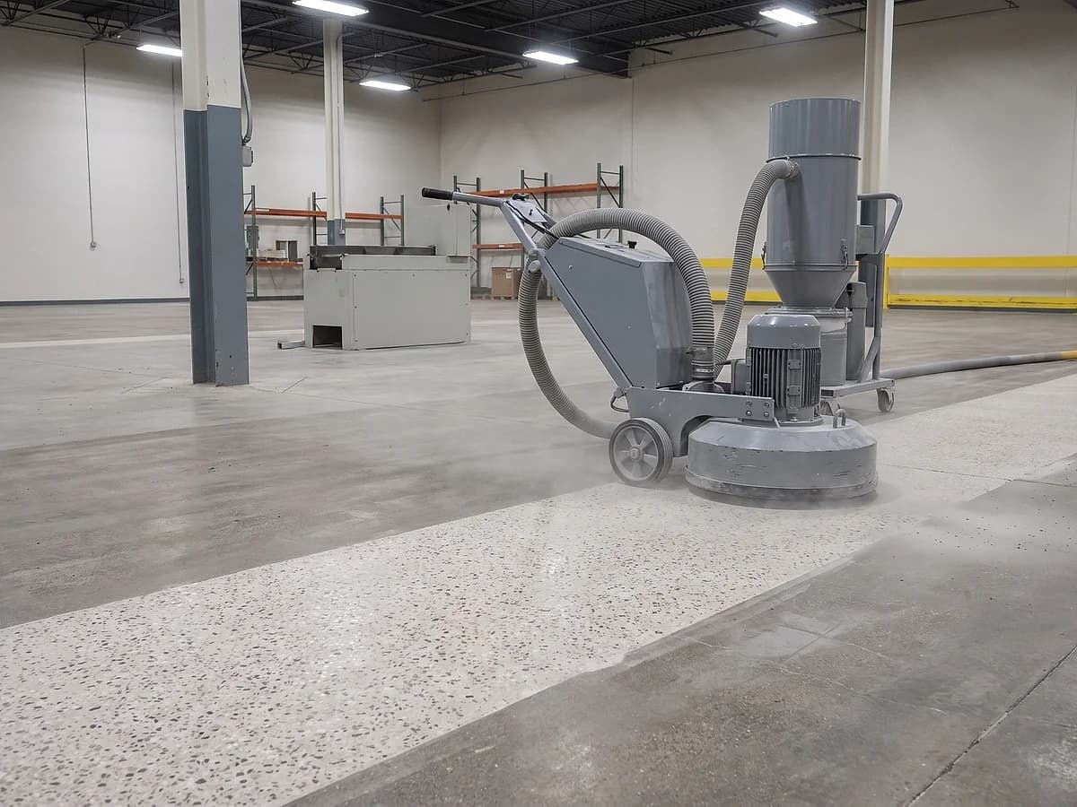 A walk-behind floor grinder with dust extraction on a plant floor, a wide band ground open in front of it exposing the aggregate