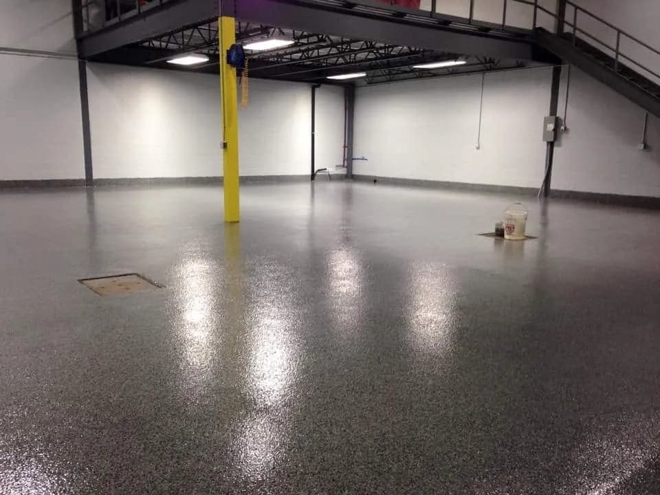 A dark grey flake floor across an industrial bay, the ceiling lights standing in it as long reflections beside a yellow column