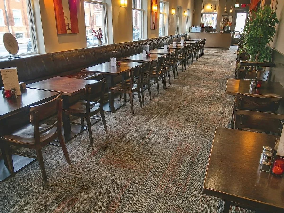 A patterned commercial carpet running through a restaurant dining room past a long banquette and a row of bare tables, laid wall to wall with the pile level and evenly cut.