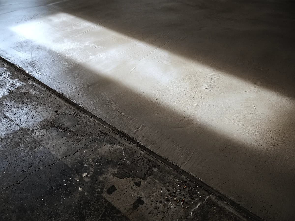 A thin cementitious overlay stopping in a feathered edge across an older slab, the new bone-grey surface beside the darker patched concrete.