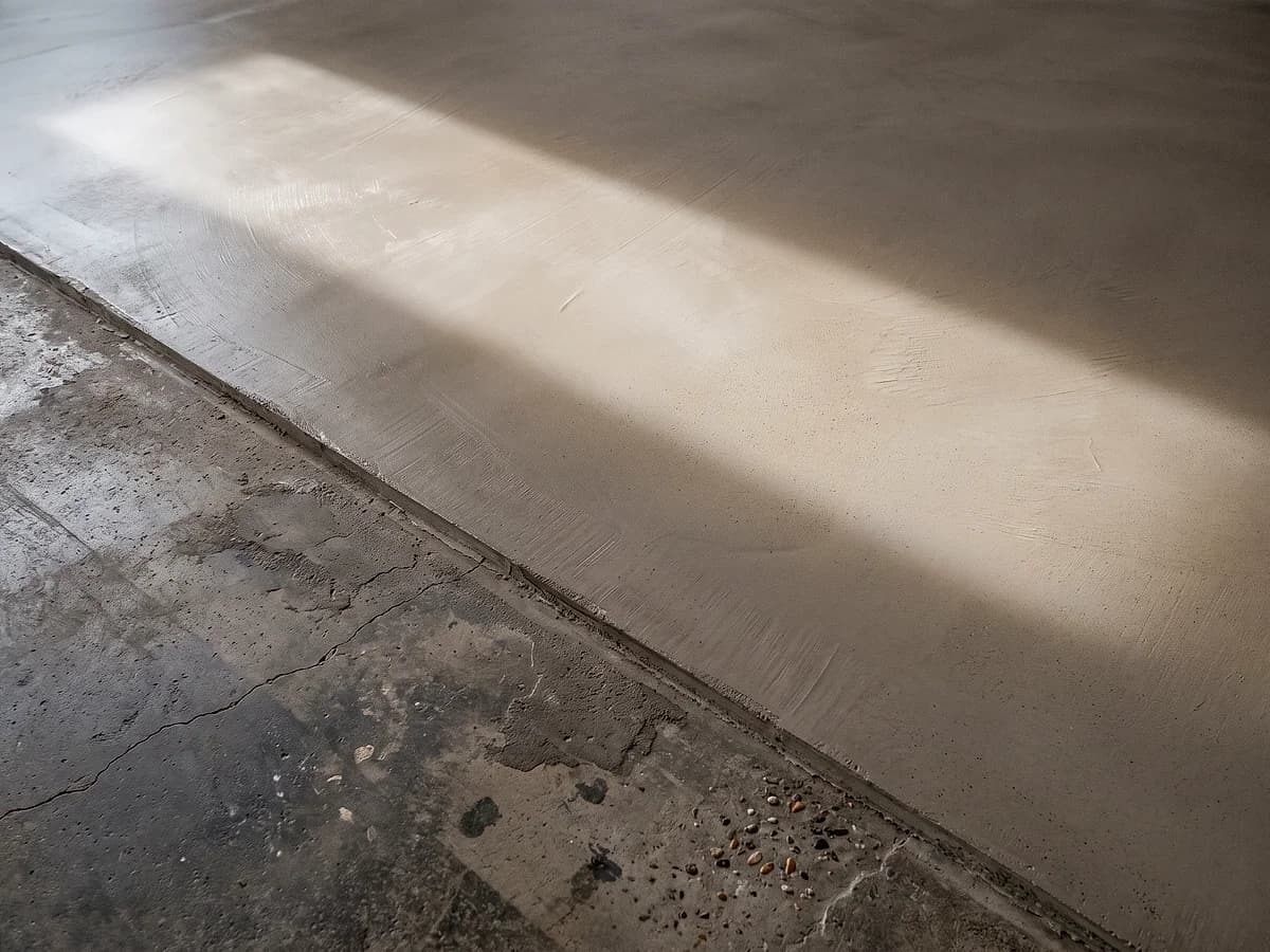A thin cementitious overlay stopping in a feathered edge across an older slab, the new bone-grey surface beside the darker patched concrete.