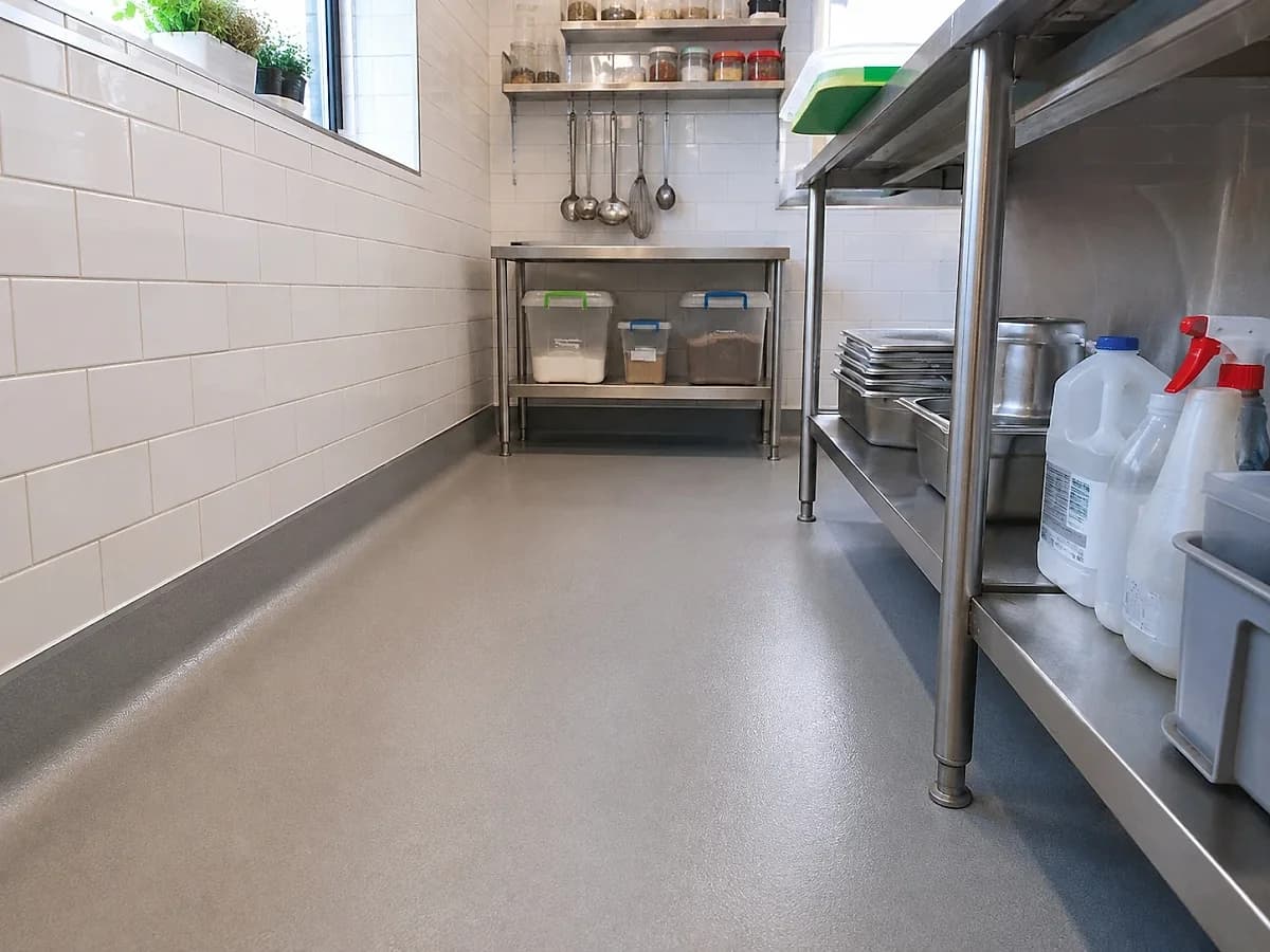 A resin floor in a commercial kitchen running up the wall in one continuous coved skirting, the same grey as the floor with no joint at the base, under a run of stainless shelving.
