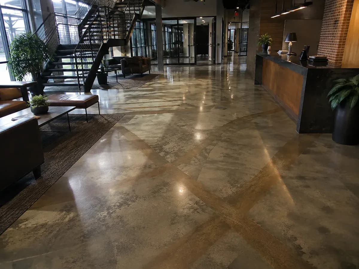 A stained concrete floor in an office building lobby, its colour clouding unevenly through warm grey tones under a satin sheen, running past a steel staircase to the reception desk.