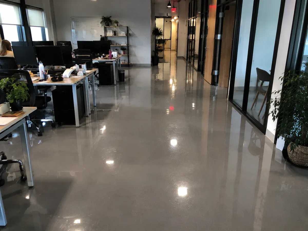 A seamless mid grey epoxy resin floor running the length of an open-plan office past a bank of desks and a run of glazed partitions, under a topcoat that holds the reflections of the ceiling lights.