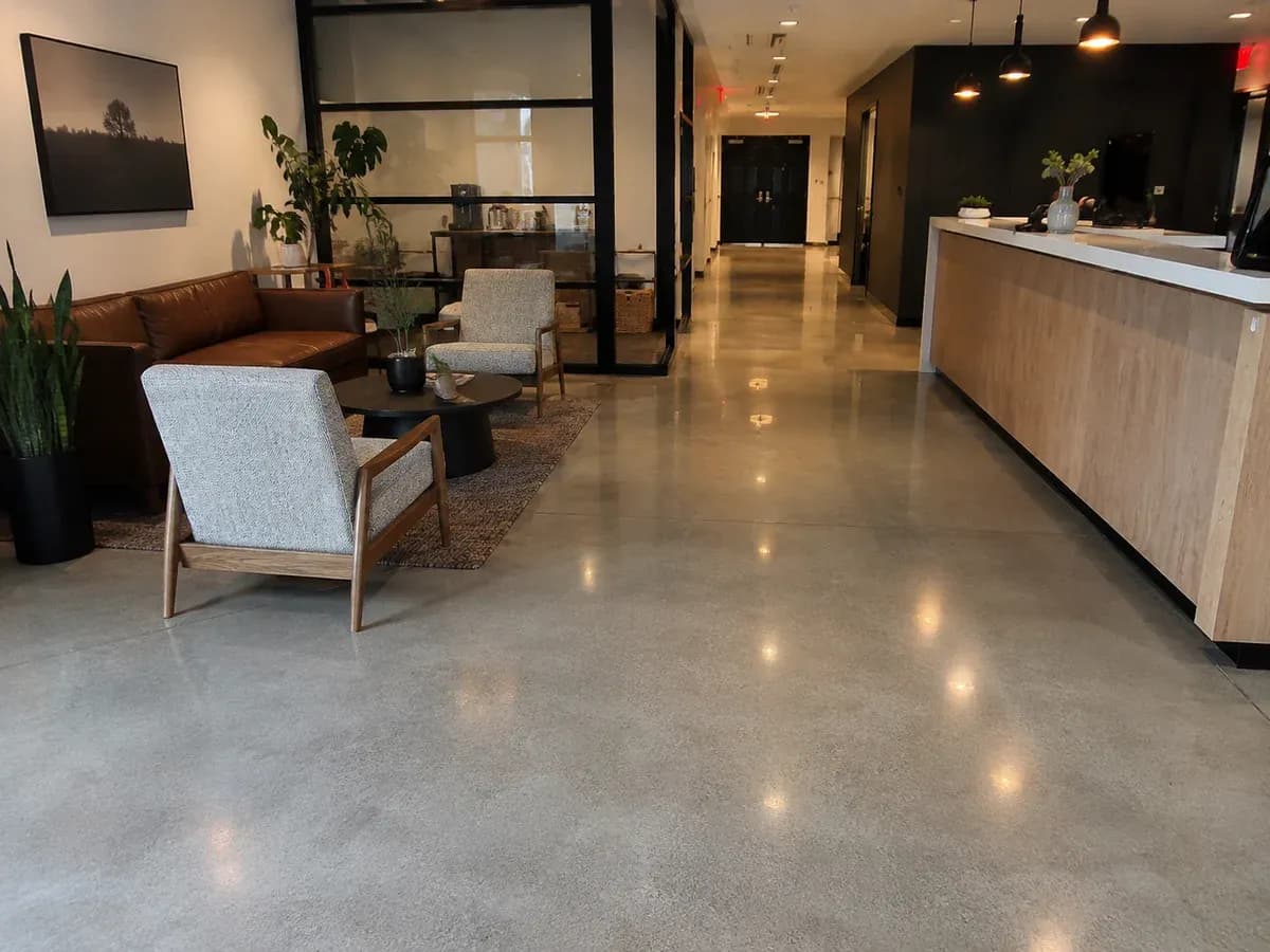 A polished concrete floor running through an office reception and lounge area past a row of soft seating to the reception counter, finished to a satin sheen that holds the reflections of the ceiling lights.