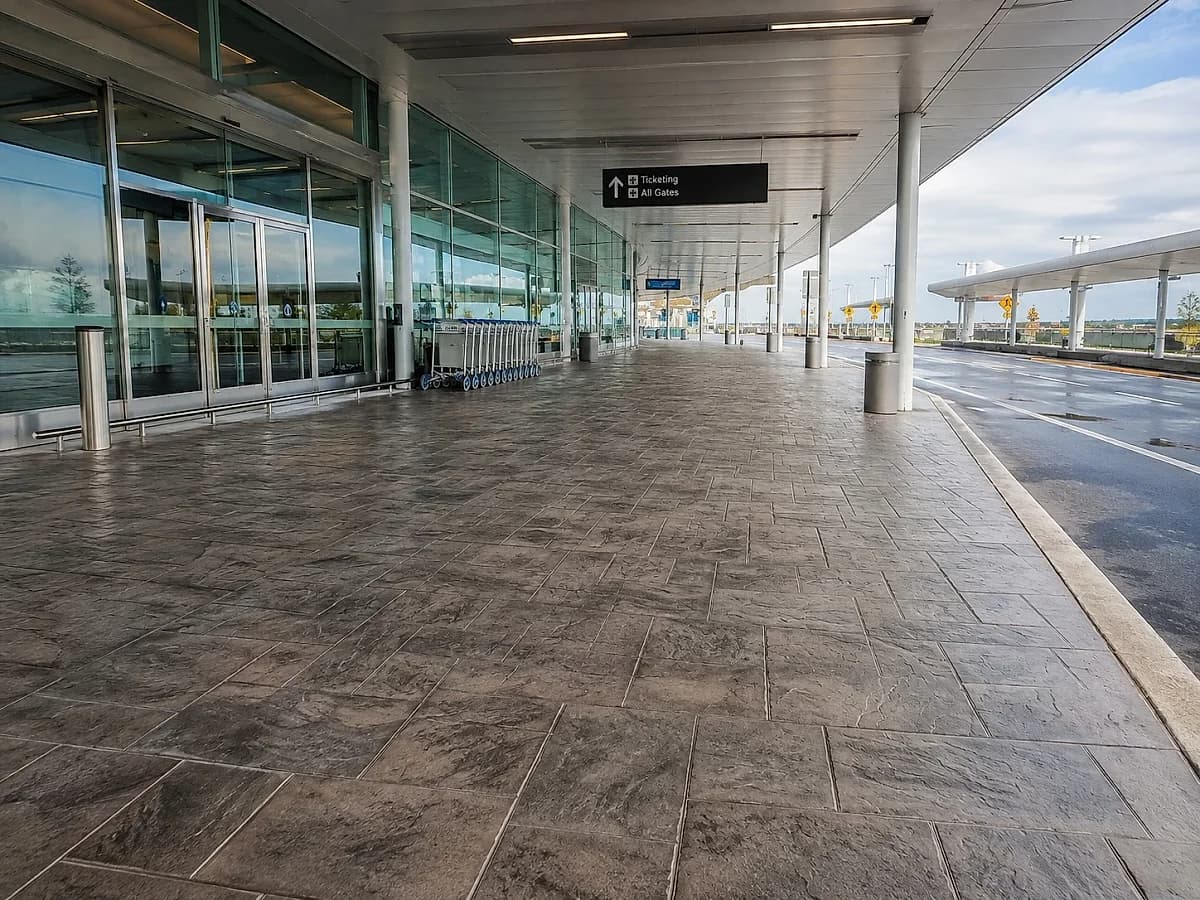 A stamped concrete walkway outside an airport terminal under the departures canopy, pressed with an irregular cut-stone pattern in mid grey with darker joint lines, running along the kerb.