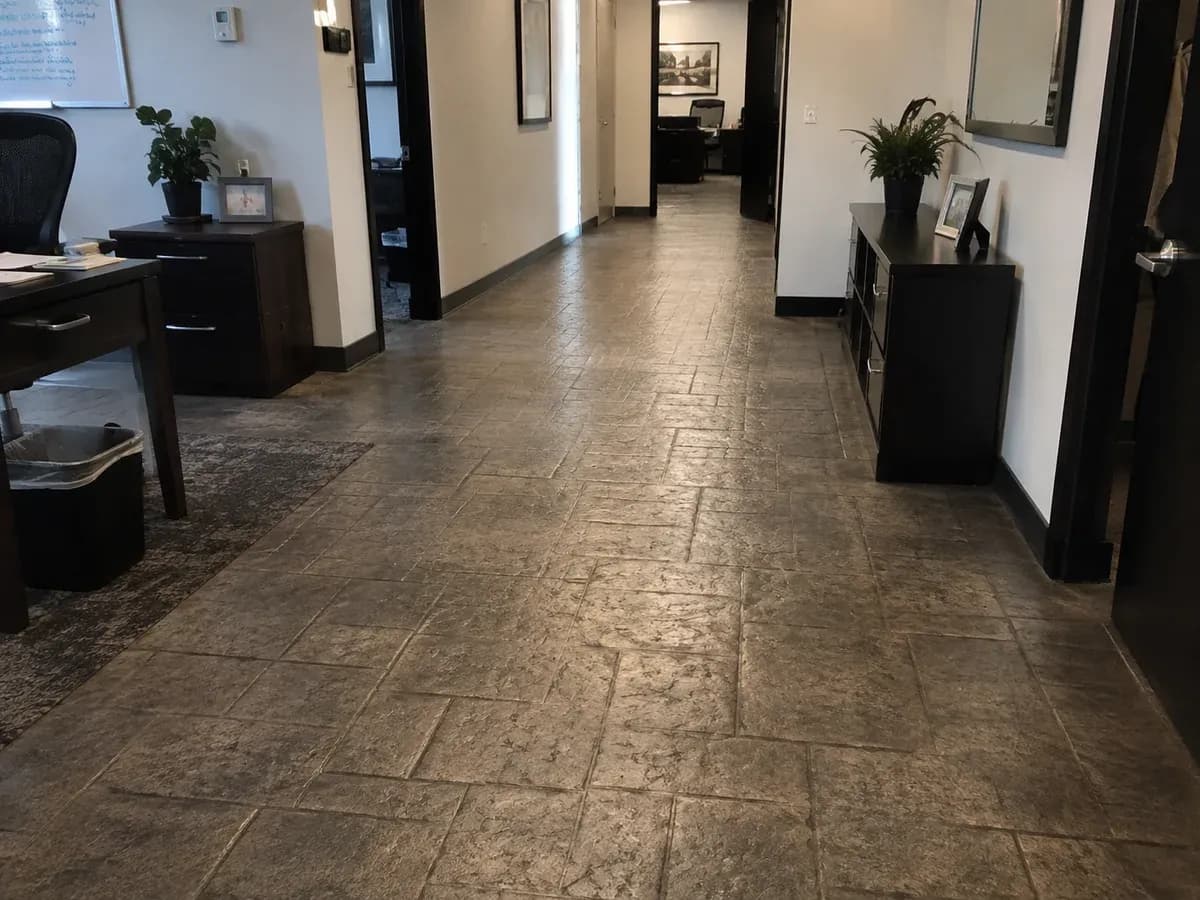 A stamped concrete floor running down an office corridor, pressed with an irregular cut-stone pattern in warm grey with darker joint lines, past a run of doors and a low cabinet.