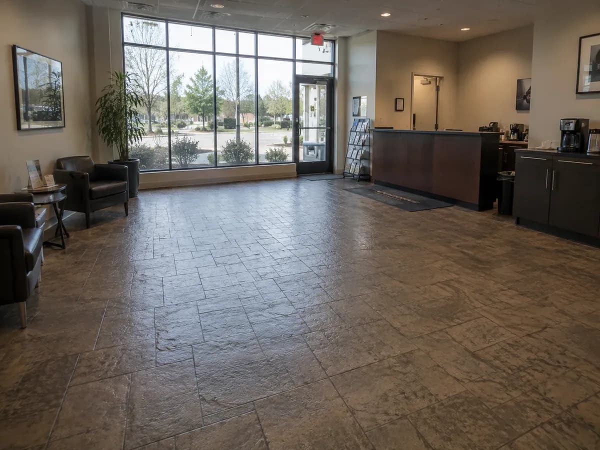 A stamped concrete floor across a showroom, pressed with a regular cut-stone pattern in warm grey with darker joint lines, running from the glazed shopfront past a seating area.