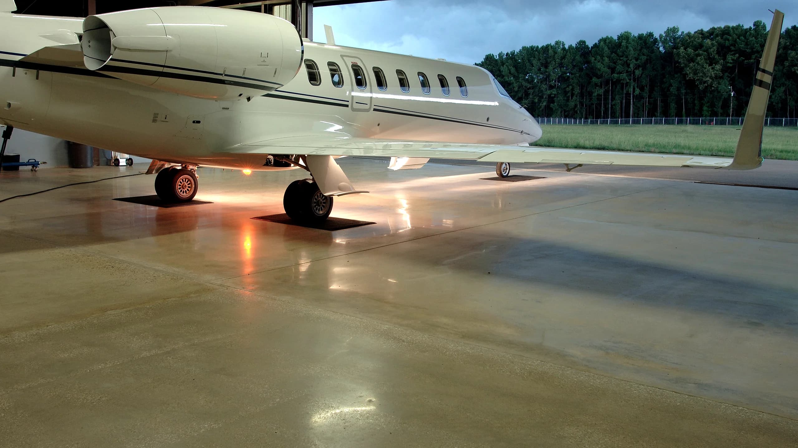 A business jet standing in a hangar with the door open onto a field at dusk, its lights running as reflections down the glossy floor