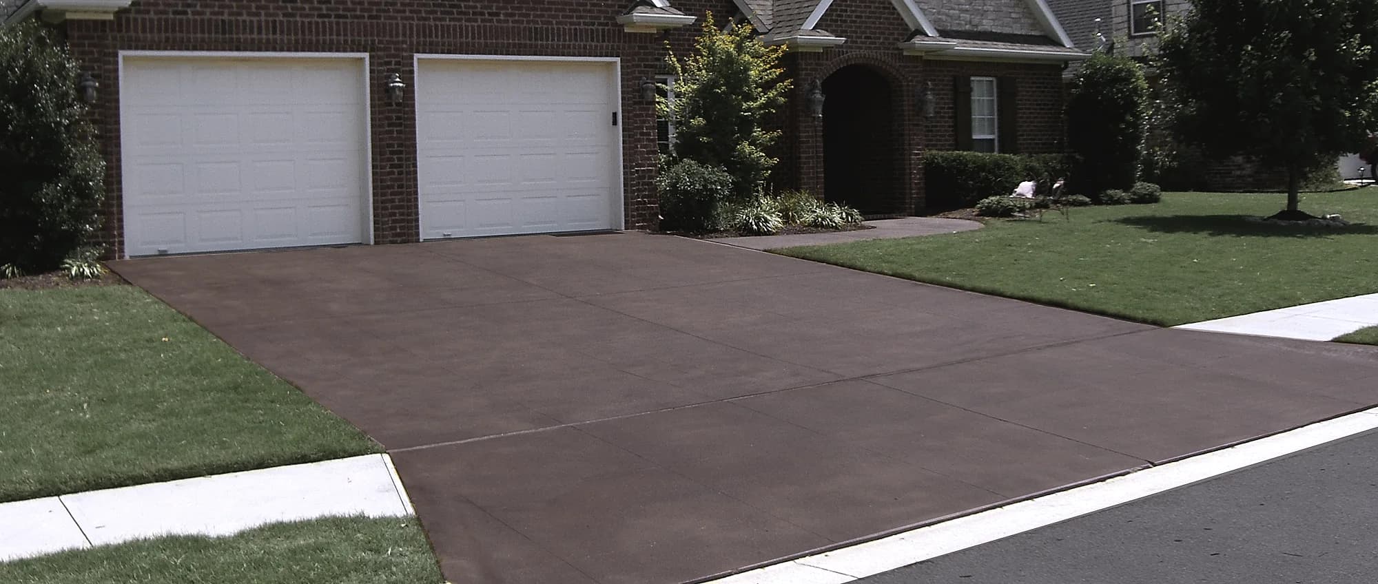 Stained and sealed concrete driveway in a warm brown tone running up to the garage doors of a brick suburban home