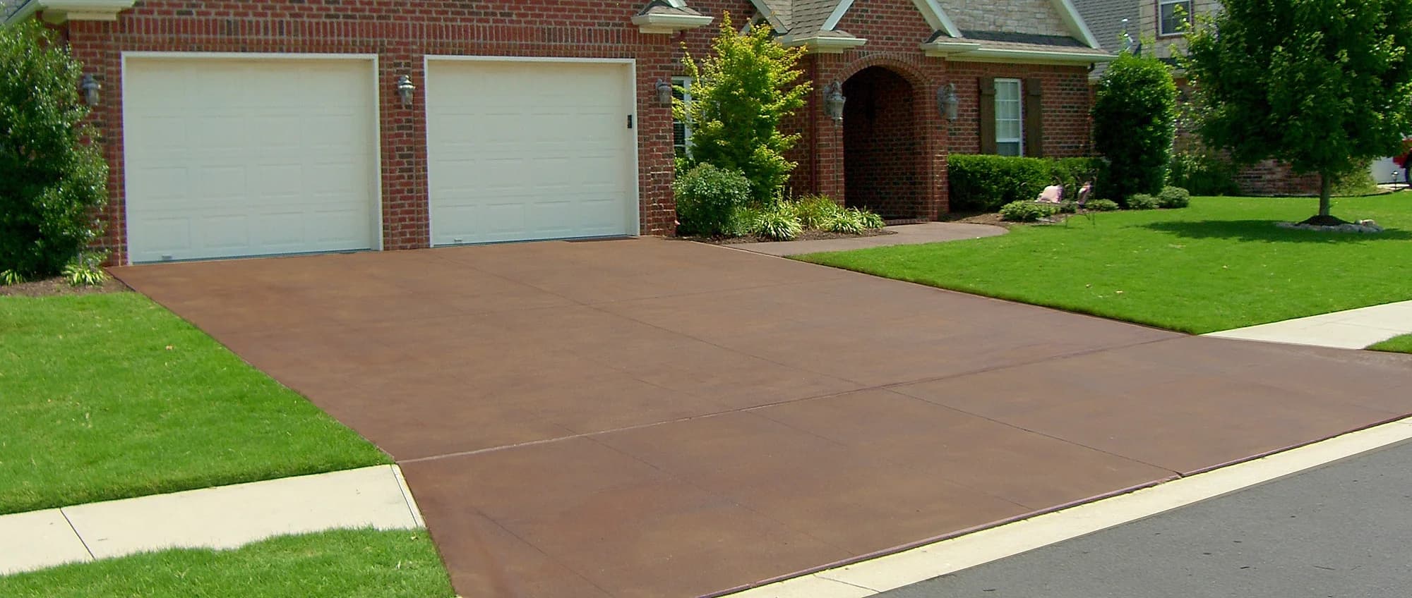 Stained and sealed concrete driveway in a warm brown tone running up to the garage doors of a brick suburban home