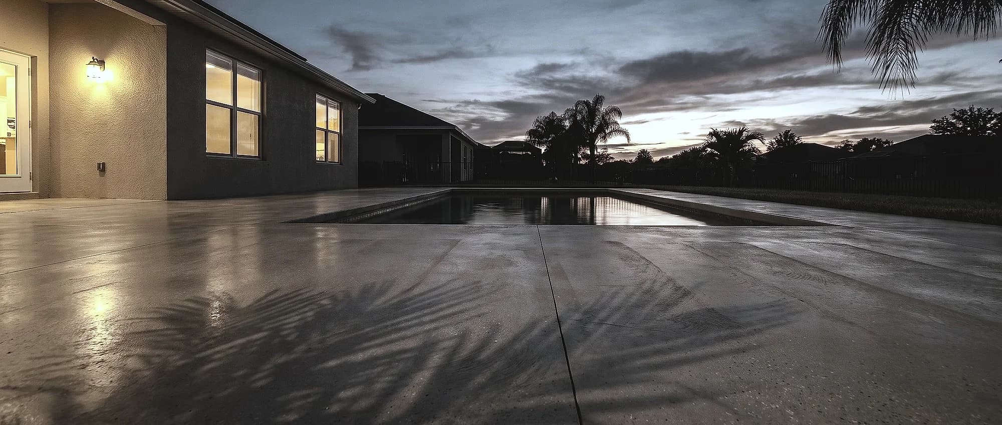 Smooth light-gray patio and pool deck at dusk with palm shadows across the surface and warm light from the house windows