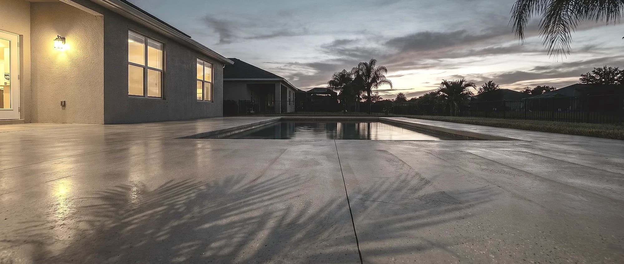 Smooth light-gray patio and pool deck at dusk with palm shadows across the surface and warm light from the house windows