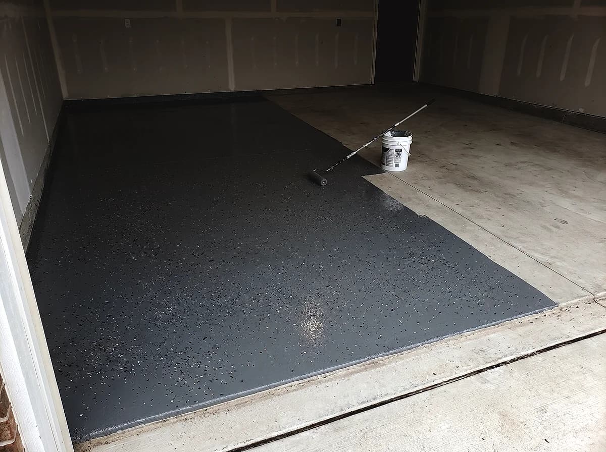 A garage floor halfway through a coating install, fresh base coat and colour flake on one half and bare concrete on the other