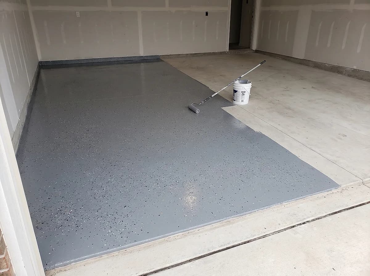 A garage floor halfway through a coating install, fresh base coat and colour flake on one half and bare concrete on the other