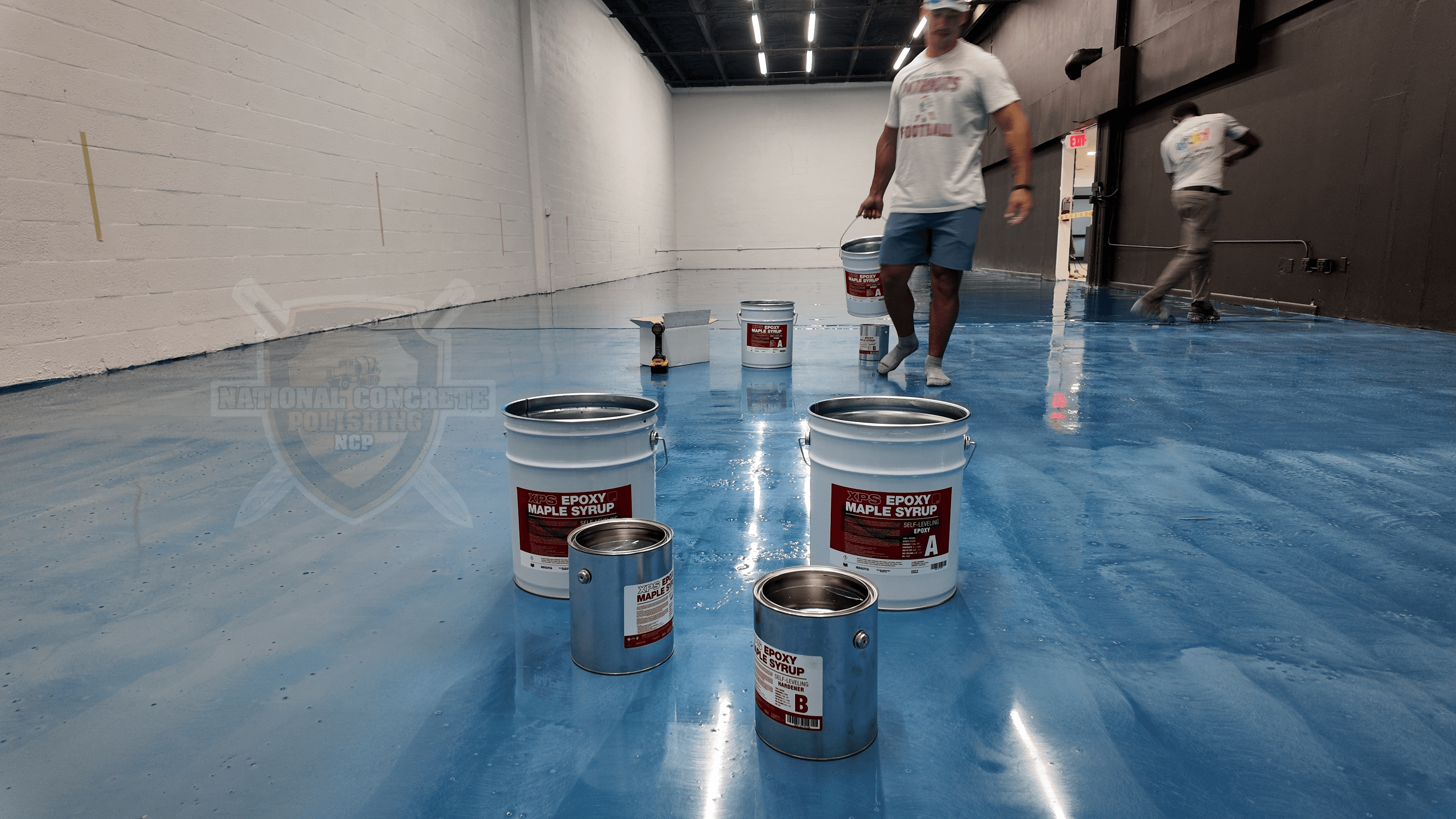 Cans of resinous coating on a metallic blue resin flooring in a warehouse setting.
