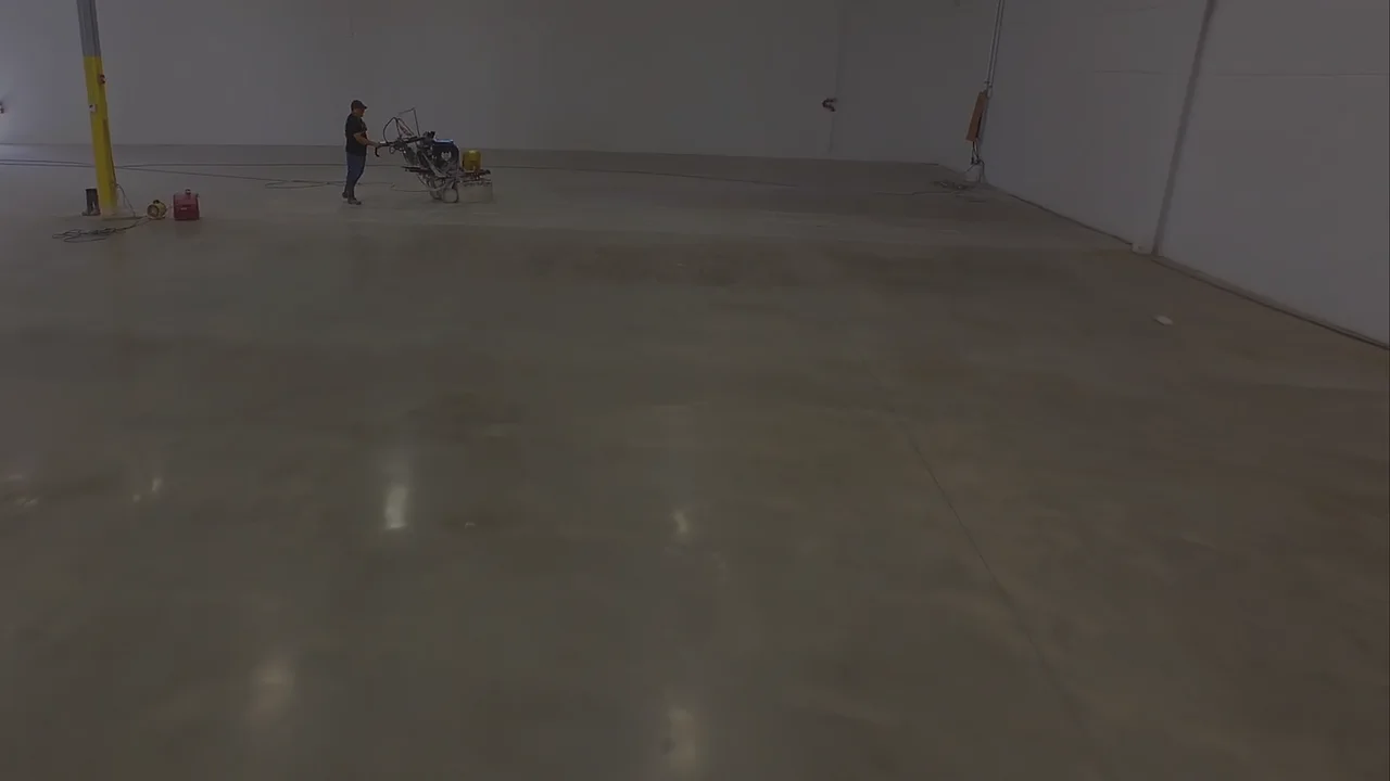 An operator steering a walk-behind grinder across a large part-polished commercial slab, white walls and a roll-up door behind him