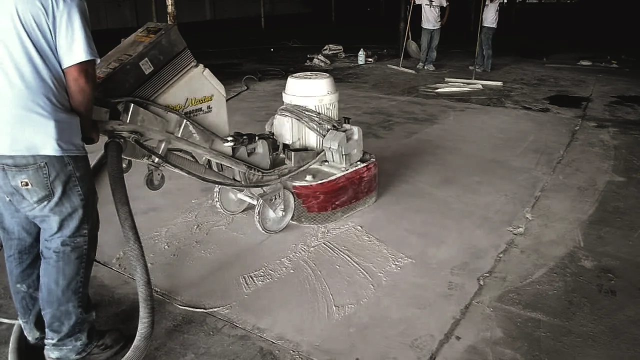 The crew working a floor grinder across a bare concrete slab, the dust extraction hose running off the machine
