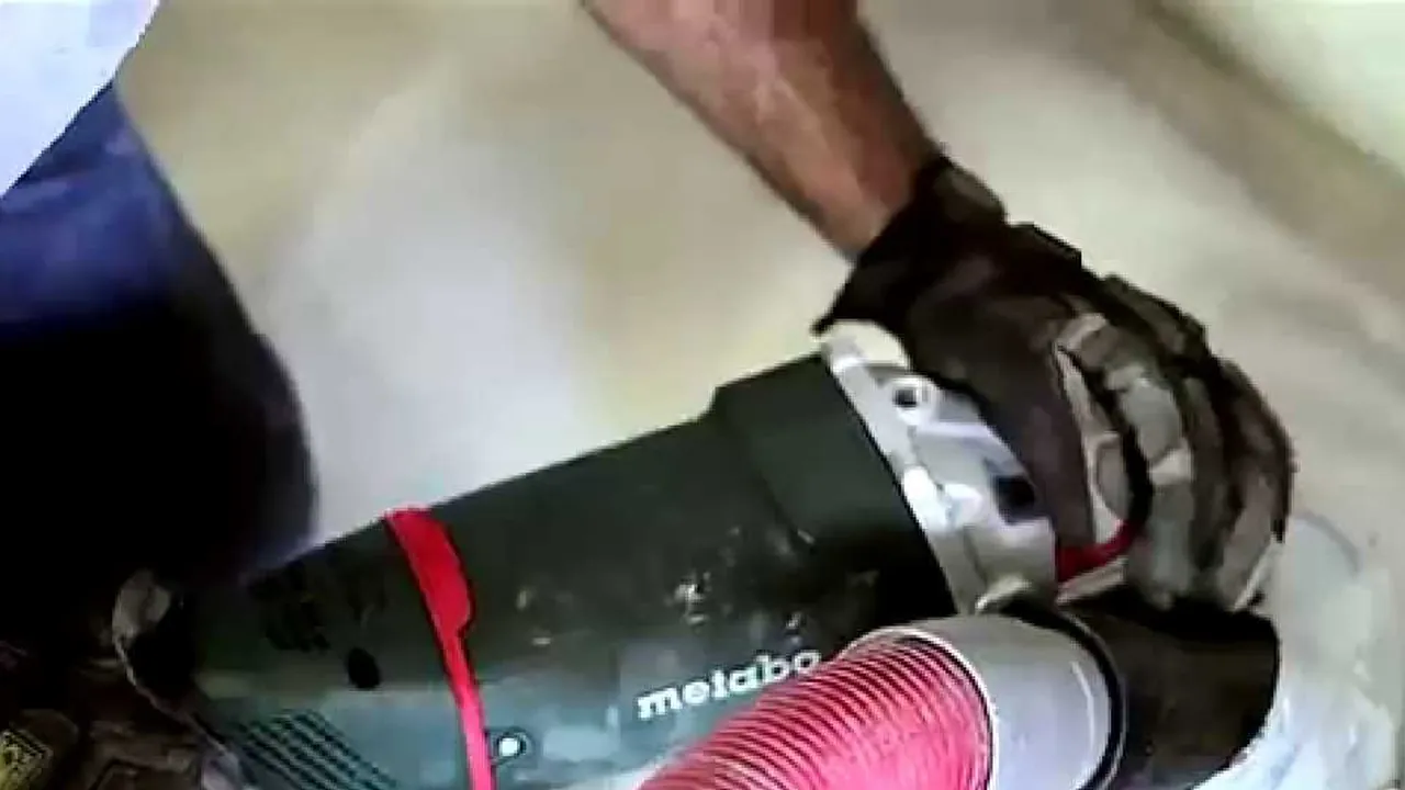 A gloved hand running a hand grinder with its dust hose along the base of a wall