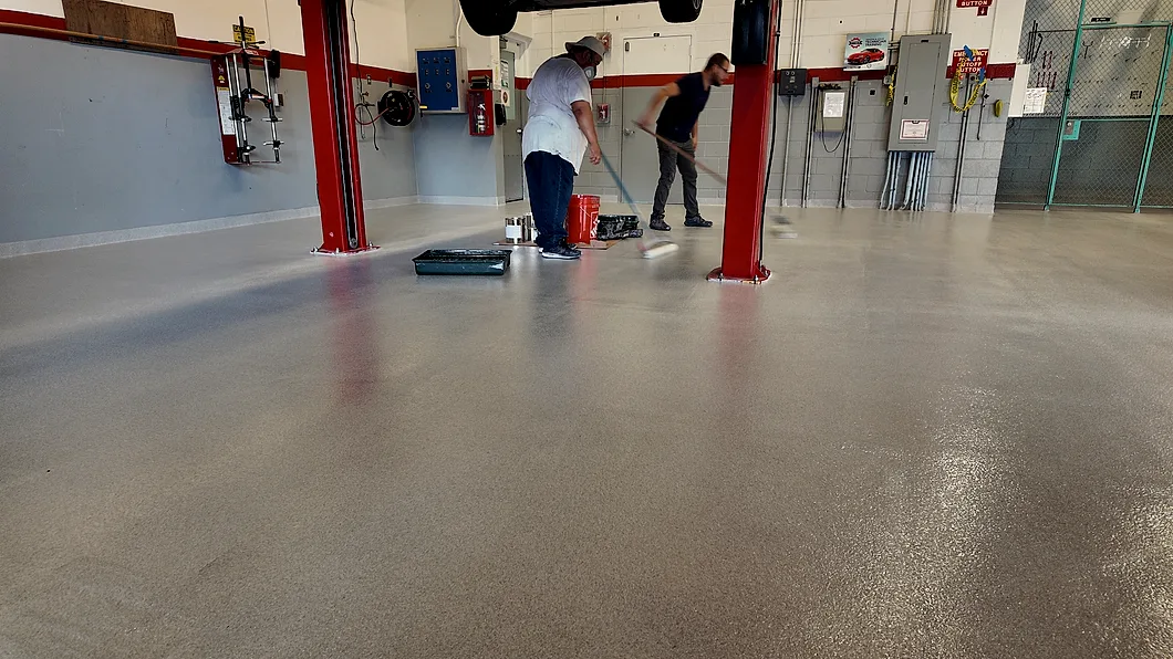 Two installers rolling the coating out across a training bay floor, one working from a roller tray beside a bucket, the other pulling a long-handled roller