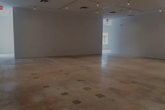 A warm polished floor with a grid of small grey patches across it, running away under gallery track lighting toward a lit doorway
