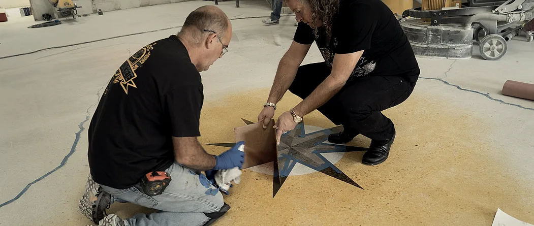 Two installers kneeling on a yellow stained floor, positioning the points of a compass star stencil with layout tape, a walk-behind floor machine standing against the wall behind them