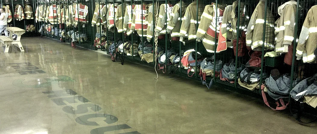Turnout gear hanging in open wire cages beside a polished bay floor, with lettering ground into the slab running away toward the far wall