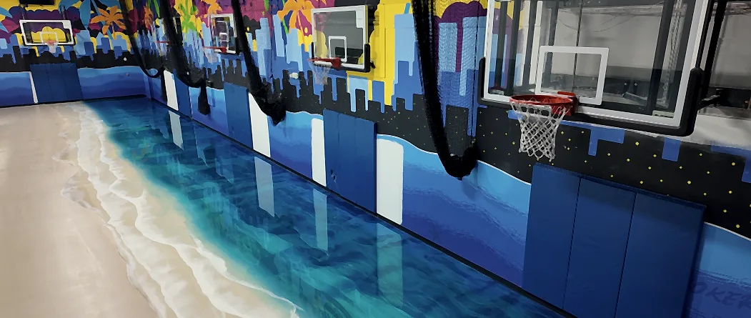 A metallic floor poured as a shoreline, sand running through white surf into deep teal water, beneath a row of backboards and painted wall padding