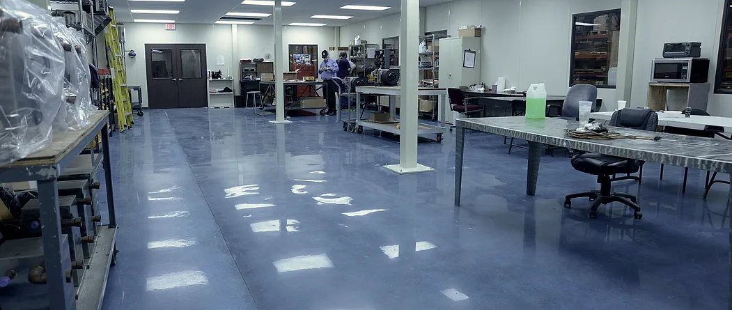 A blue stained polished floor running the length of a working assembly room, past benches, parts trolleys and two people at work