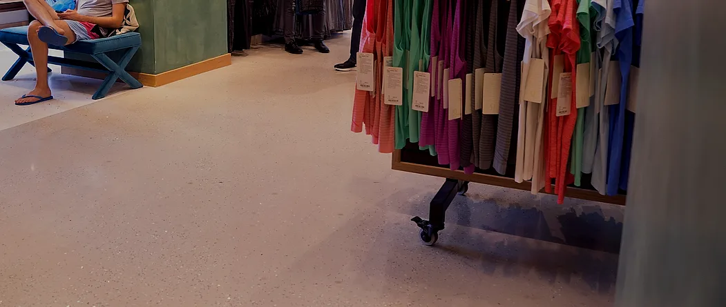 A pale terrazzo-look floor with fine glass aggregate running across a store during trading hours, past rolling racks and a seating bench