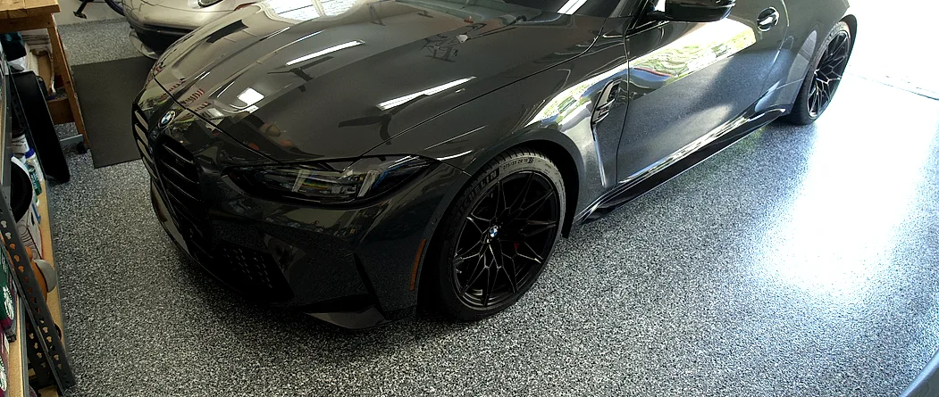 A gray and black flake floor filling a home garage, with a coupe parked across it, shelving along the wall and daylight coming in through the open door