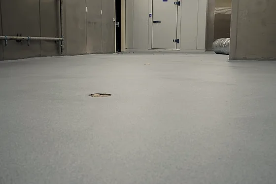 A pale grey quartz floor seen low across the kitchen toward the walk-in doors, a floor drain set into it