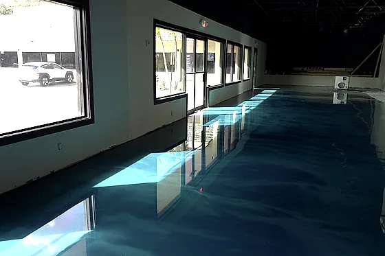 A turquoise metallic floor running the length of an empty room along a row of street windows, daylight laid across it in bright panes