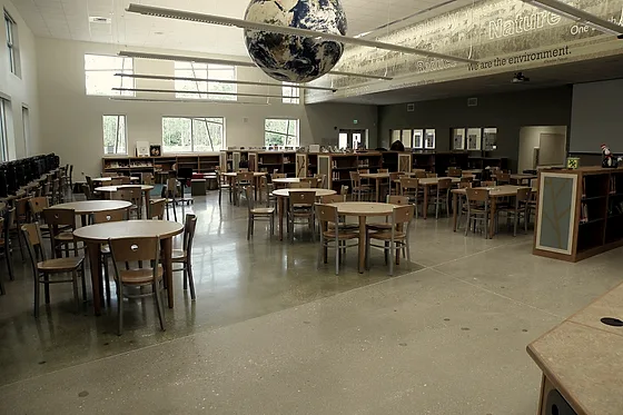 A furnished cafeteria and library over a pale polished floor, round tables and low shelving standing on it under a high glazed wall